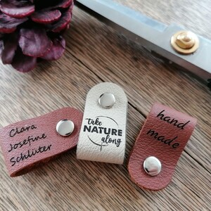 May include: Three leather tags with silver snaps. One is light beige with "take NATURE along" printed on it. Another is brown with "hand made" printed on it. The third is brown with a name printed on it. Scissors and a pinecone are in the background.
