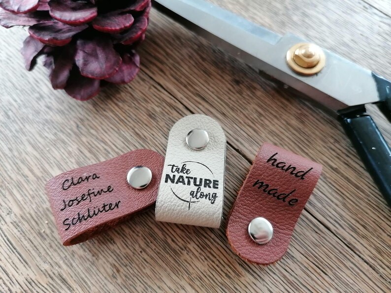 May include: Three leather tags with silver snaps. One is light beige with "take NATURE along" printed on it. Another is brown with "hand made" printed on it. The third is brown with a name printed on it. Scissors and a pinecone are in the background.