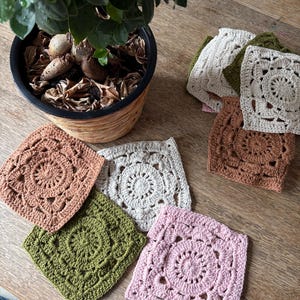 May include: A collection of square crochet motifs featuring intricate circular patterns in the center. The set includes various colors such as dusty pink, olive green, cream, and terracotta, arranged in a stack and scattered on a wooden surface.