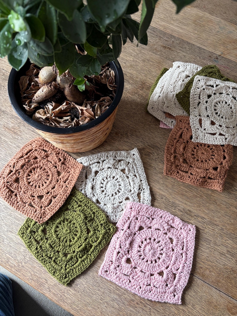 May include: A collection of square crochet motifs featuring intricate circular patterns in the center. The set includes various colors such as dusty pink, olive green, cream, and terracotta, arranged in a stack and scattered on a wooden surface.