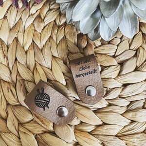May include: Two brown leather tags with silver snaps. One tag features a yarn ball and needles graphic. The other tag has the text "mit viel Liebe hergestellt" embossed on it. The tags are on a woven, light brown surface.