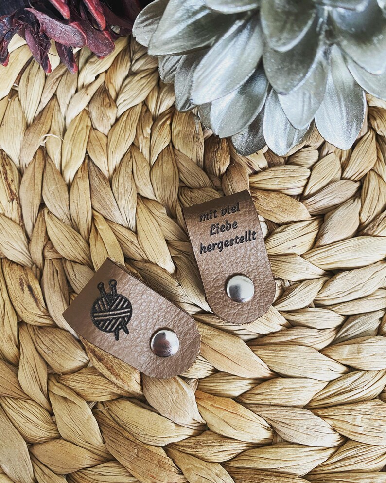 May include: Two brown leather tags with silver snaps. One tag features a yarn ball and needles graphic. The other tag has the text "mit viel Liebe hergestellt" embossed on it. The tags are on a woven, light brown surface.