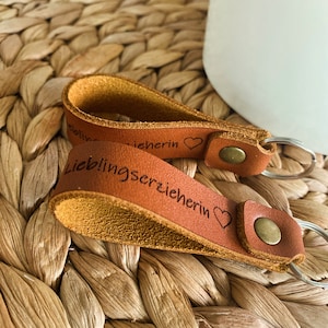 May include: Two brown leather keychains with metal rings. The keychain on top has the text "Liebherin" with a heart symbol. The keychain on the bottom has the text "Lieblingserzieherin" with a heart symbol.