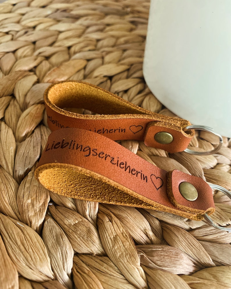 May include: Two brown leather keychains with metal rings. The keychain on top has the text "Liebherin" with a heart symbol. The keychain on the bottom has the text "Lieblingserzieherin" with a heart symbol.