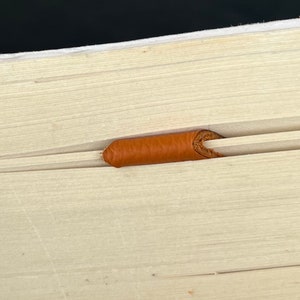 Peut inclure: Une surface en bois clair pr&eacute;sentant une rainure horizontale avec un petit insert cylindrique en cuir orange-brun plac&eacute; au centre du canal. Le grain du bois est visible et la pi&egrave;ce en cuir pr&eacute;sente une texture lisse et arrondie.