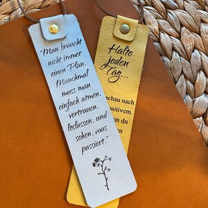 May include: Two white leather bookmarks with yellow leather accents. One bookmark has the text "Man braucht nicht immer einen Plan. Manchmal muss man einfach atmen, vertrauen, loslassen, und sehen, was passiert." The other bookmark has the text "Halte jeden Tag... schau nach positivem."