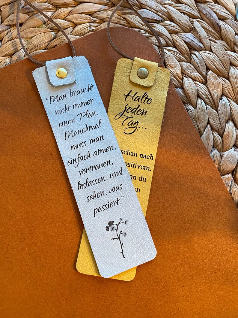 May include: Two white leather bookmarks with yellow leather accents. One bookmark has the text "Man braucht nicht immer einen Plan. Manchmal muss man einfach atmen, vertrauen, loslassen, und sehen, was passiert." The other bookmark has the text "Halte jeden Tag... schau nach positivem."