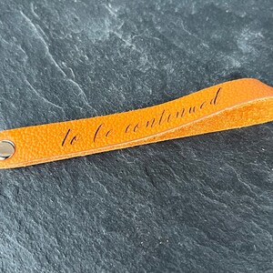 Peut inclure: Un marque-page en cuir orange vif avec une surface textur&eacute;e et un rivet en m&eacute;tal argent&eacute; &agrave; une extr&eacute;mit&eacute;. La phrase "to be continued" est imprim&eacute;e en &eacute;criture cursive noire le long du centre de la bande de cuir orange.