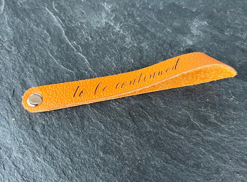 Peut inclure: Un marque-page en cuir orange vif avec une surface textur&eacute;e et un rivet en m&eacute;tal argent&eacute; &agrave; une extr&eacute;mit&eacute;. La phrase "to be continued" est imprim&eacute;e en &eacute;criture cursive noire le long du centre de la bande de cuir orange.