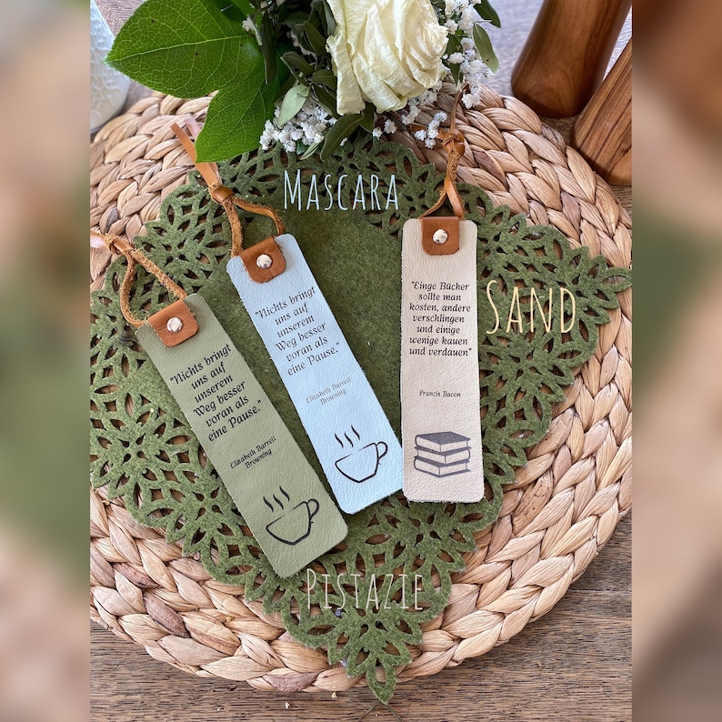 May include: Three leather bookmarks with different colors and designs. The bookmarks feature quotes about reading and include the words "Mascara", "Sand", and "Pistazie".