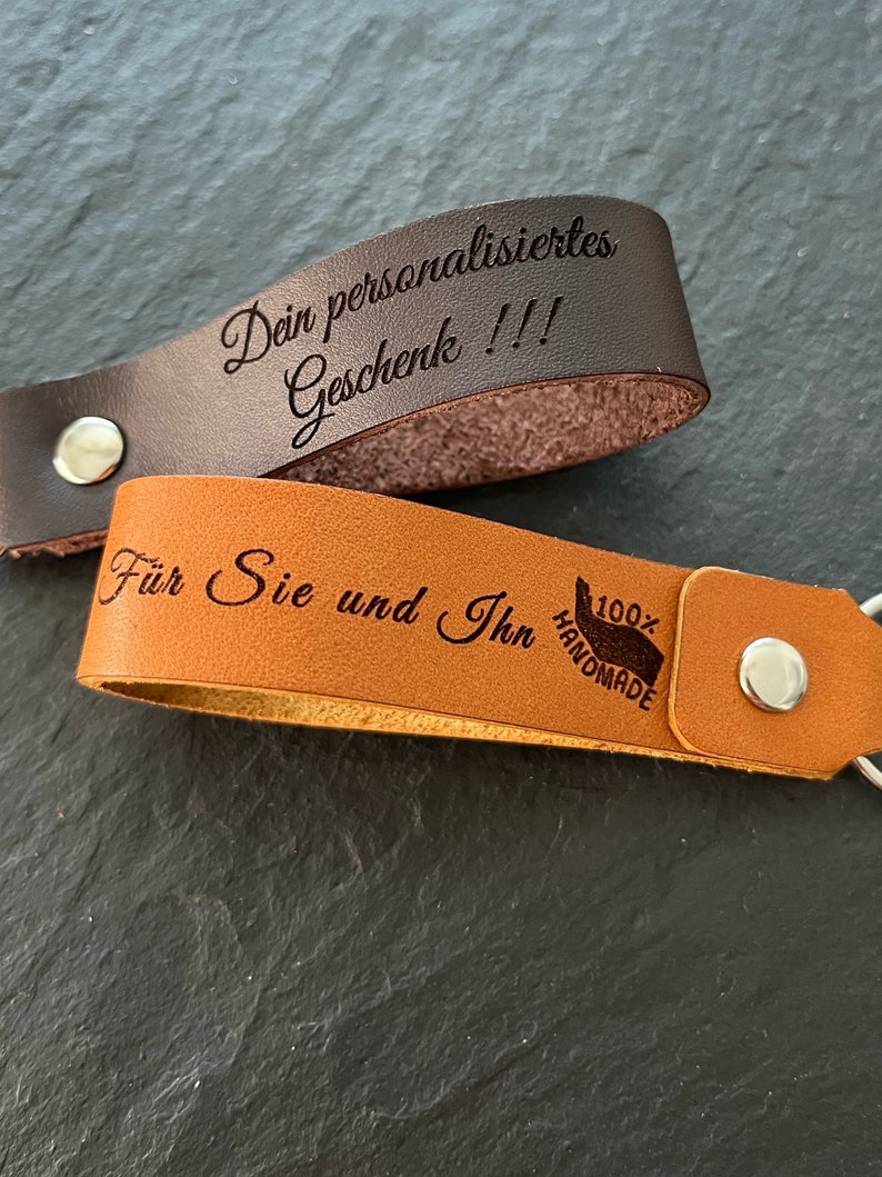 May include: Two leather keychains with engraved text. The top keychain is brown and says "Dein personalisiertes Geschenk!!!". The bottom keychain is tan and says "F&uuml;r Sie und Ihn 100% HANDMADE".