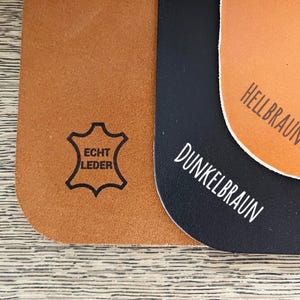 Op de afbeelding: Drie lederen swatches in bruin, donkerbruin en zwart. De bruine swatch heeft de tekst "Echt Leder" erop gedrukt. De donkerbruine swatch heeft de tekst "Dunkelbraun" erop gedrukt. De zwarte swatch heeft de tekst "Hellbraun" erop gedrukt.