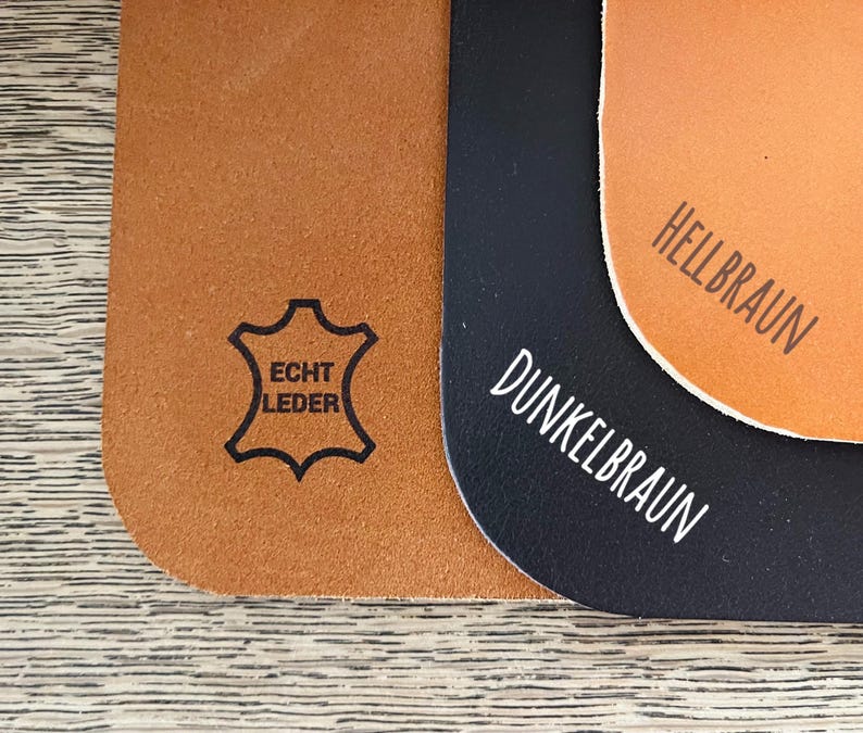 Op de afbeelding: Drie lederen swatches in bruin, donkerbruin en zwart. De bruine swatch heeft de tekst "Echt Leder" erop gedrukt. De donkerbruine swatch heeft de tekst "Dunkelbraun" erop gedrukt. De zwarte swatch heeft de tekst "Hellbraun" erop gedrukt.