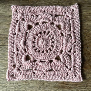 May include: A square crochet motif in a soft pastel pink yarn. The design features a circular center with radiating stitches, transitioning into a square shape with openwork shell patterns and decorative gaps throughout the textured fabric.