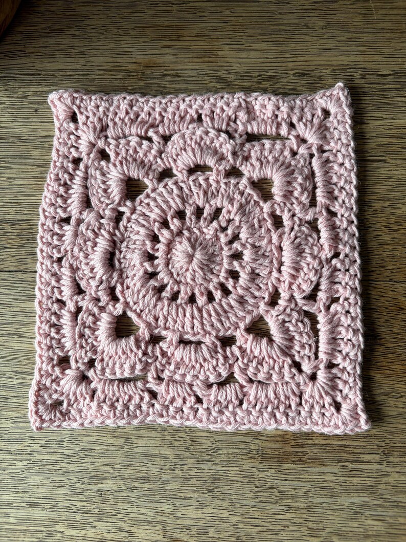 May include: A square crochet motif in a soft pastel pink yarn. The design features a circular center with radiating stitches, transitioning into a square shape with openwork shell patterns and decorative gaps throughout the textured fabric.