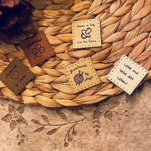 May include: Five small, square, brown leather patches with different designs and text. The patches include a four-leaf clover, a pacifier, a yarn ball and knitting needles, a heart, and a branch with leaves. The text on the patches reads "Gl&uuml;ckskind", "Gimme a hug I'm the future", "Handmade by Anja", "Knitted with by Christine", and "Lebe und liebe das Leben!"