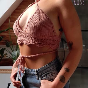 Peut inclure: Haut de bikini en crochet rose poudr&eacute; avec bord festonn&eacute; et fermeture &agrave; nouer. Le haut est fait &agrave; la main et pr&eacute;sente un col en V. Le mot "ROSE" est visible en arri&egrave;re-plan. Il est associ&eacute; &agrave; un jean en denim bleu.