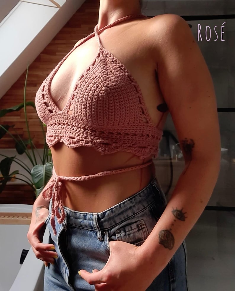 Peut inclure: Haut de bikini en crochet rose poudr&eacute; avec bord festonn&eacute; et fermeture &agrave; nouer. Le haut est fait &agrave; la main et pr&eacute;sente un col en V. Le mot "ROSE" est visible en arri&egrave;re-plan. Il est associ&eacute; &agrave; un jean en denim bleu.