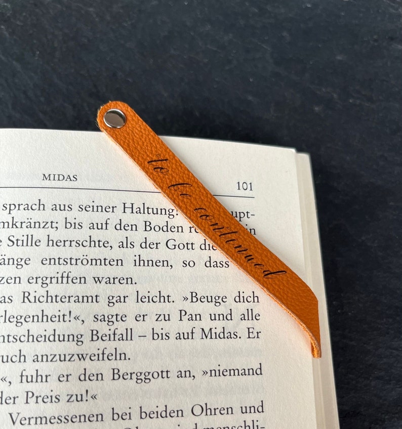 Peut inclure: Un marque-page en cuir orange textur&eacute; avec un rivet m&eacute;tallique argent&eacute; sur le dessus et le texte cursif noir to be continued imprim&eacute; en diagonale sur toute la longueur de la bande, pos&eacute; sur une page de livre ouverte avec du texte imprim&eacute;.