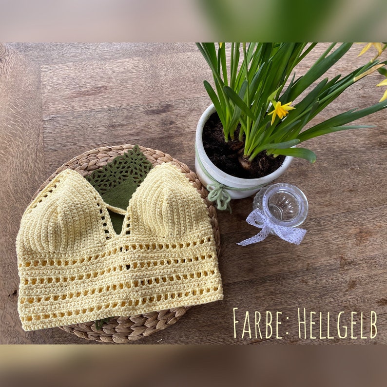 May include: A crocheted crop top in a light yellow color. The top has a V-neckline and a criss-cross design in the front. The text "FARBE: HELLGELB" is visible in the bottom right corner of the image.