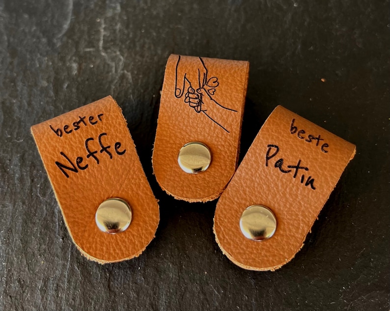 May include: Three brown leather tags with silver snaps. One tag reads "bester Neffe", another features a hand-holding illustration, and the third says "beste Patin". The tags are on a dark surface.