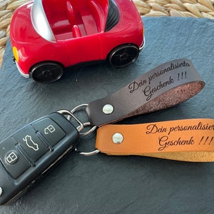 May include: A red toy car with a black key chain attached. The key chain has two leather tags with the text "Dein personalisiertes Geschenk!!!" and "Dein personalisiertes Geschenk!!!"
