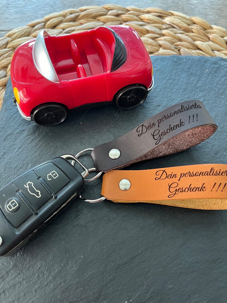 May include: A red toy car with a black key chain attached. The key chain has two leather tags with the text "Dein personalisiertes Geschenk!!!" and "Dein personalisiertes Geschenk!!!"