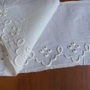 Könnte beinhalten: Weißer Stoff mit gestickten Blumen und einem wellenförmigen Rand. Der Stoff ist gefaltet, wodurch die Textur und das Design sichtbar werden. Die Stickerei weist kleine, kreisförmige Akzente auf, die ein zartes und verziertes Muster bilden. Dieser Stoff eignet sich für verschiedene Bastelprojekte.