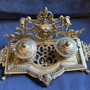 May include: Ornate antique brass inkwell with two ink pots and a decorative lion's head. The inkwell features intricate floral and scrollwork designs, with a rectangular base and small feet. A vintage desk accessory.