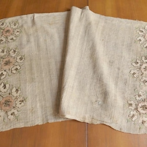May include: Beige table runner with floral embroidery in pink, white, and green. The runner has fringed edges and is made of a textured fabric. The floral design is repeated on both ends of the runner.