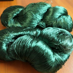 Può includere: Due matasse di filato verde smeraldo sono esposte su una superficie di legno. Il filato è ben avvolto, con una consistenza lucida e setosa. L'illuminazione evidenzia la profondità del colore e i fili sottili del filato.