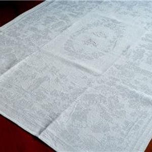 Puede incluir: Mantel de damasco blanco con un estampado floral. El mantel está doblado, con una sección cuadrada más pequeña en la parte superior. La tela tiene un brillo sutil y el diseño es visible en el tejido.