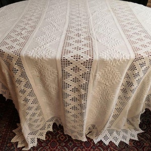 Pode incluir: Uma toalha de mesa branca de crochê com um delicado padrão de renda. A toalha de mesa está colocada sobre uma mesa redonda de madeira.