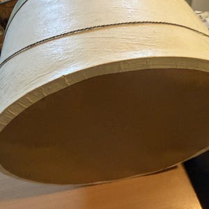 May include: A large, cylindrical hat box with a cream-colored exterior and a brown interior. The box is accented with a thin, dark rope detail around the middle and a light green trim around the top.
