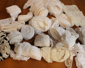 Op de afbeelding: Een assortiment witte en crèmekleurige kanten linten en versieringen, waaronder opgerolde spoelen en gevouwen stukken. De verschillende texturen en patronen zijn zichtbaar, met enkele stukken met open ontwerpen. De items zijn gerangschikt op een houten oppervlak.