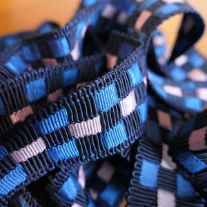 May include: Close-up of a navy blue ribbon with a woven pattern of blue and light pink squares. The ribbon has a textured, fringed edge. The ribbon is likely used for sewing, crafting, or decorative purposes.