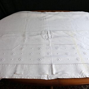 May include: White linen tablecloth with embroidered floral details and a monogrammed initial "H" in the center.