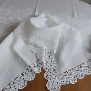 May include: White linen tablecloth with delicate floral embroidery and scalloped lace trim. The tablecloth is folded, revealing the intricate details of the lace and the subtle pattern of the embroidery.