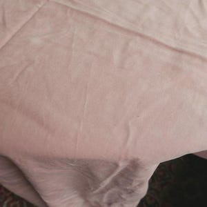 Op de afbeelding: Een close-up van een lichtroze stof met zichtbare plooien en kreukels. De stof lijkt zacht, mogelijk zijde of linnen. De belichting creëert schaduwen die de textuur van het materiaal benadrukken.