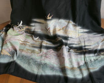 May include: A black garment featuring a detailed landscape print with flying cranes, stylized mountains, and pine trees in shades of white, pale green, and soft pink. The fabric displays a flowing, layered composition with dark shadows and light highlights.