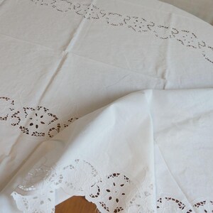 Pode incluir: Toalha de mesa branca com intrincados desenhos florais vazados nas bordas. O tecido está dobrado, mostrando a textura e os detalhes do trabalho vazado. Adequado para uma mesa de jantar ou uma ocasião especial.