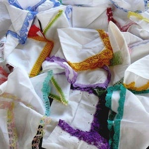 May include: A pile of white handkerchiefs with decorative lace borders. The lace is in a variety of colours, such as blue, yellow, purple, green, and pink. The handkerchiefs are folded and arranged, creating a textured appearance.