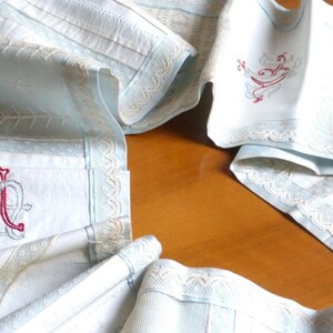 May include: Four white linen table runners with blue and cream lace trim. The runners have embroidered designs in red thread. One runner has the letters "A" and "M" intertwined, another has a bird design, and the other two have floral designs.