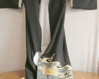 May include: A black kimono featuring a white inner lining and wide sleeves. The lower portion of the garment is decorated with a stylised wave pattern in shades of light blue, gold, and white. Two small circular white emblems are visible on the upper chest.