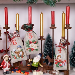 Op de afbeelding: Een traditionele adventskalender met zes stoffen zakjes, elk met een ander nummer geborduurd erop. De zakjes hangen aan een metalen standaard met zes kandelaars. De kaarsen zijn rood en goud. Er zijn ook kleine kerstversieringen op de standaard.