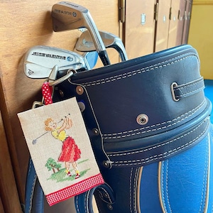 May include: A blue and white golf bag with a brown leather strap. A small embroidered tag with a red, white, and green design of a woman playing golf is attached to the bag. The tag says "Jessie Valentine" on it.