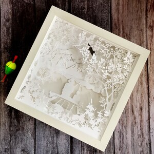 Op de afbeelding: Witte papier-uitkniparbeid in een wit frame. De kunst toont een bos sc&egrave;ne met bomen, bladeren en een klein gebouw in de verte.