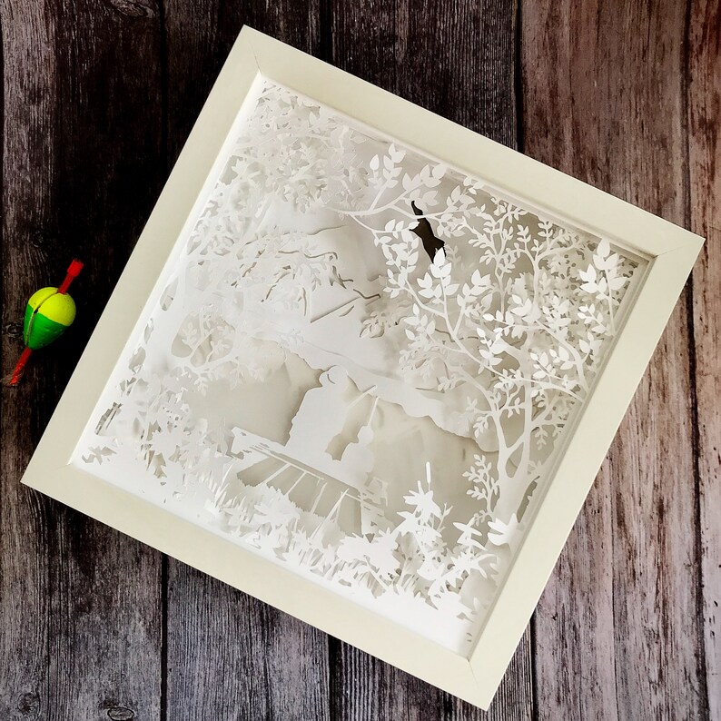 Op de afbeelding: Witte papier-uitkniparbeid in een wit frame. De kunst toont een bos sc&egrave;ne met bomen, bladeren en een klein gebouw in de verte.