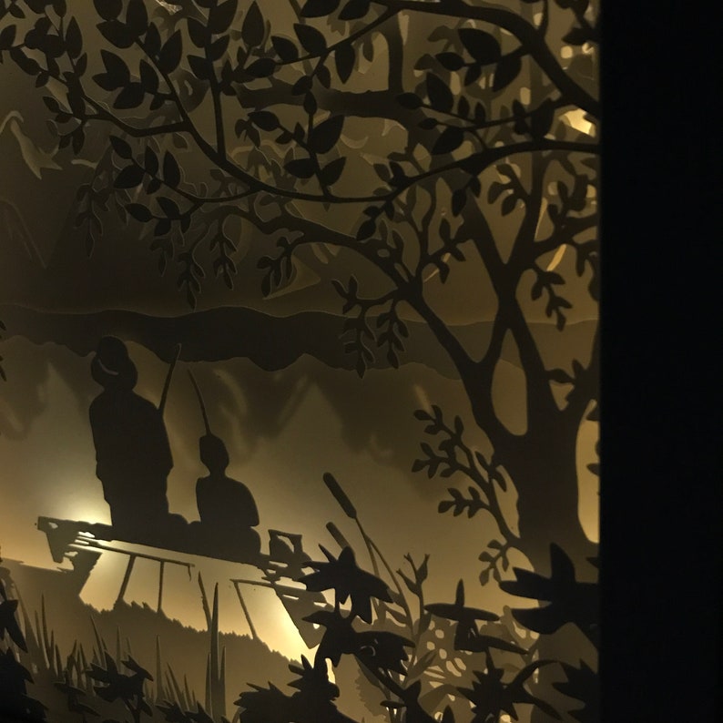 Op de afbeelding: Een kunstwerk van papier snijwerk dat twee figuren toont die op een platform in een bossetting zitten. De figuren zijn gesilhouetteerd tegen een lichte achtergrond, en het bos is gemaakt van ingewikkelde papier gesneden bomen en bladeren.