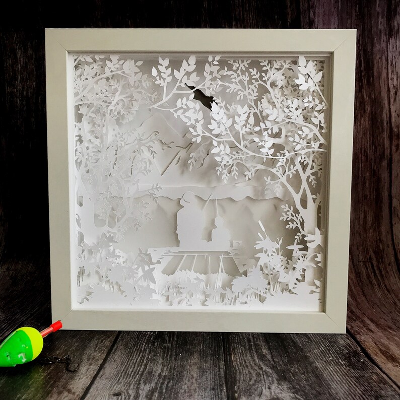 Op de afbeelding: Een wit papierkunstwerk in een wit frame dat een sc&egrave;ne voorstelt van een persoon die aan het vissen is in een meer met bomen en bergen op de achtergrond.